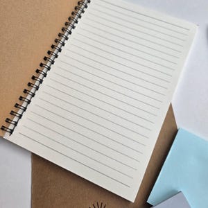May include: An open spiral-bound notebook with lined pages, resting on a brown cover. The notebook's pages are white with black lines. The brown cover has the text "chlo&CO CREATES" printed in black. Several pastel-colored sticky notes are in the background.