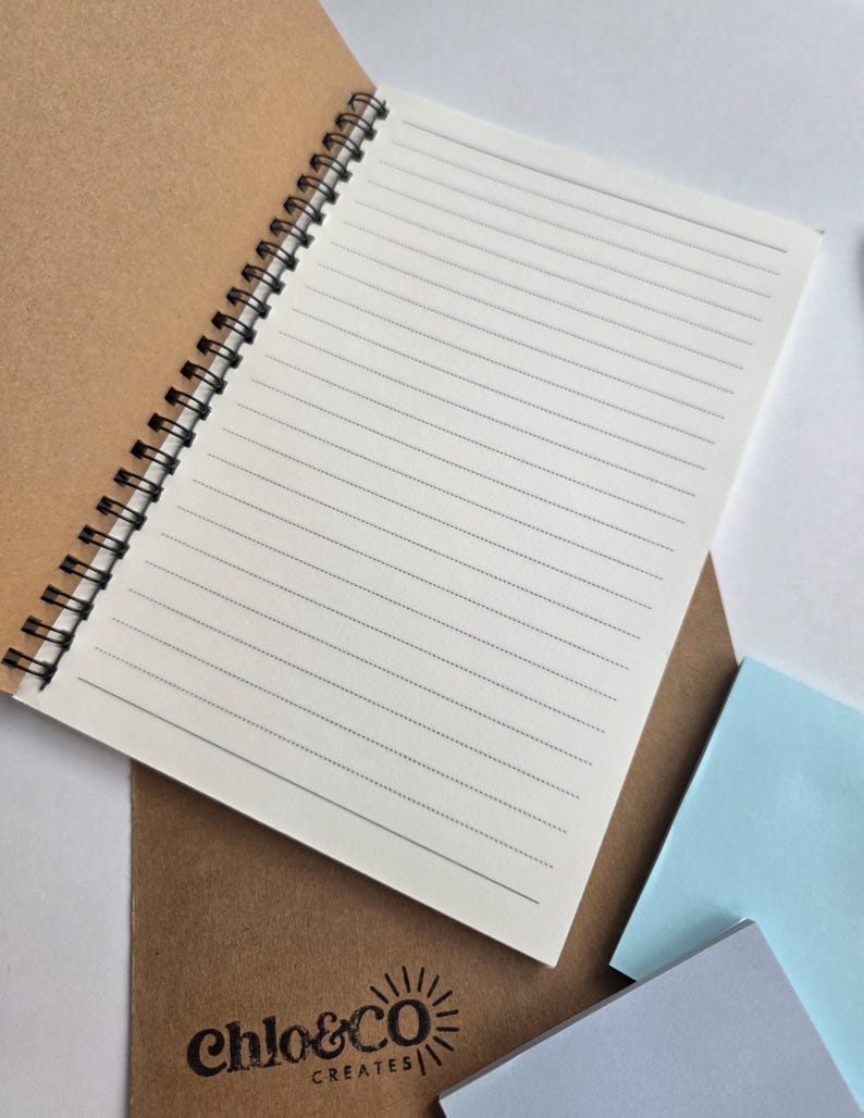 May include: An open spiral-bound notebook with lined pages, resting on a brown cover. The notebook's pages are white with black lines. The brown cover has the text "chlo&CO CREATES" printed in black. Several pastel-colored sticky notes are in the background.