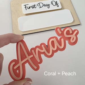 May include: A coral and peach coloured acrylic name sign that says "Aria's". The sign is a popular choice for a first day of school or a birthday.