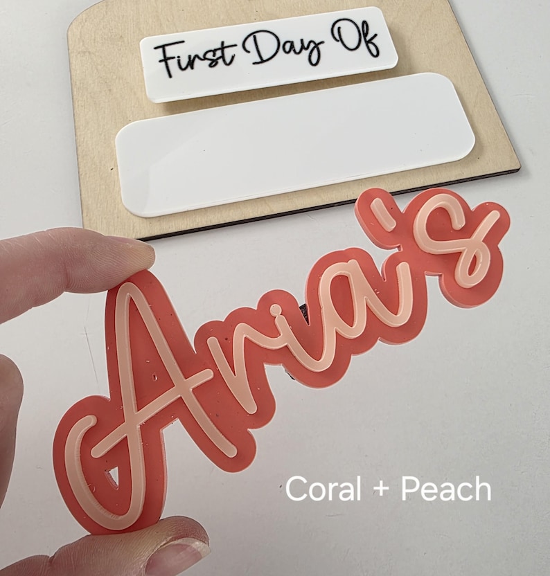 May include: A coral and peach coloured acrylic name sign that says "Aria's". The sign is a popular choice for a first day of school or a birthday.