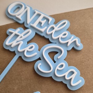 May include: A light blue and white cake topper featuring the layered text "ONE der the Sea" in a combination of block and script fonts. The design consists of a light blue backing layer with white raised lettering on top, attached to a thin stem.