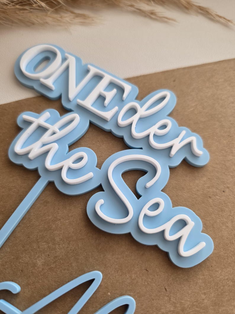 May include: A light blue and white cake topper featuring the layered text "ONE der the Sea" in a combination of block and script fonts. The design consists of a light blue backing layer with white raised lettering on top, attached to a thin stem.