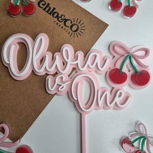 May include: Pink acrylic cake topper with the words "Olivia is One" in white script. The topper features a pink bow and two red cherries.