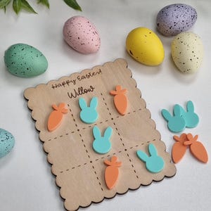 May include: A wooden game board with a scalloped edge. The board has a grid pattern with nine squares. The squares are filled with images of carrots and bunnies. The board is labeled "Happy Easter! Willow". There are also several colorful Easter eggs and bunny and carrot game pieces scattered around the board.