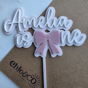 May include: White and pink acrylic cake topper with the text "Amelia is one" and a pink bow. The topper is on a brown background.