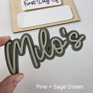 May include: Sage green acrylic name sign with the name "Milo's" in a cursive font. The sign is on a white background with the text "First Day Of" on a white rectangle.