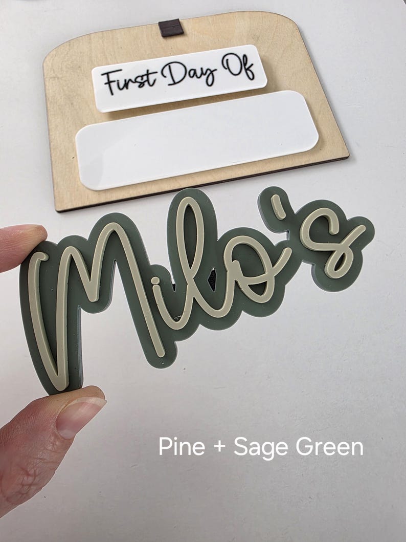 May include: Sage green acrylic name sign with the name "Milo's" in a cursive font. The sign is on a white background with the text "First Day Of" on a white rectangle.