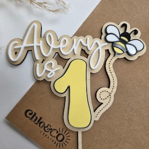 Peut inclure: Décoration de gâteau en bois avec le nom "Avery" en écriture blanche, les mots "is 1" en jaune et noir, et une illustration d'abeille. La décoration est sur une carte marron avec le logo "chloe&co creates".