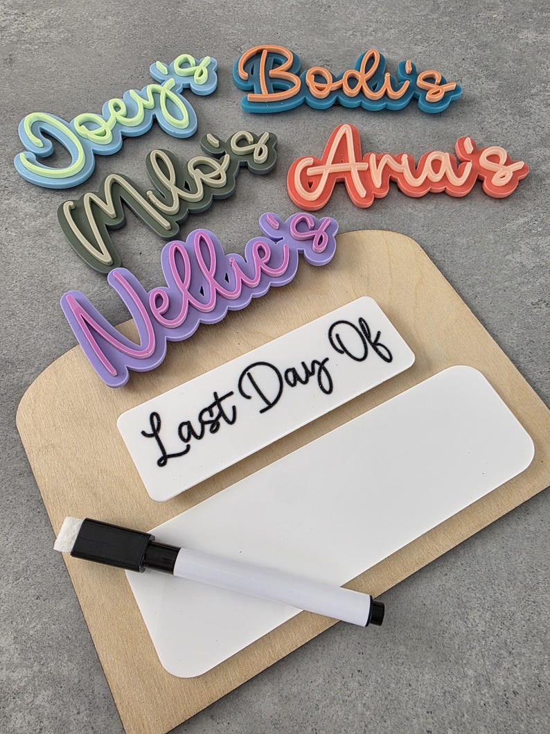 May include: A wooden board with a white dry-erase surface and a black marker. The board has a sign that says "Last Day Of" and several colourful, 3D, plastic name tags that say "Joey's", "Bodi's", "Milo's", "Aria's", and "Nellie's".