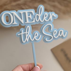 May include: A light blue and white cake topper featuring the text ONEder the Sea in a layered design. The topper consists of a light blue backing with white raised lettering attached to a thin light blue vertical stick for insertion into a cake.