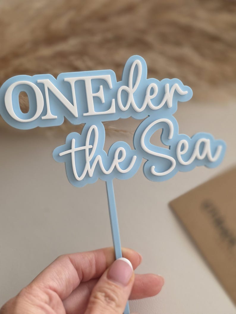 May include: A light blue and white cake topper featuring the text ONEder the Sea in a layered design. The topper consists of a light blue backing with white raised lettering attached to a thin light blue vertical stick for insertion into a cake.