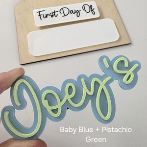May include: A light blue and pistachio green "Joe's" name sign with a cursive font. The sign is made of wood and has a blank space for a date or other text.
