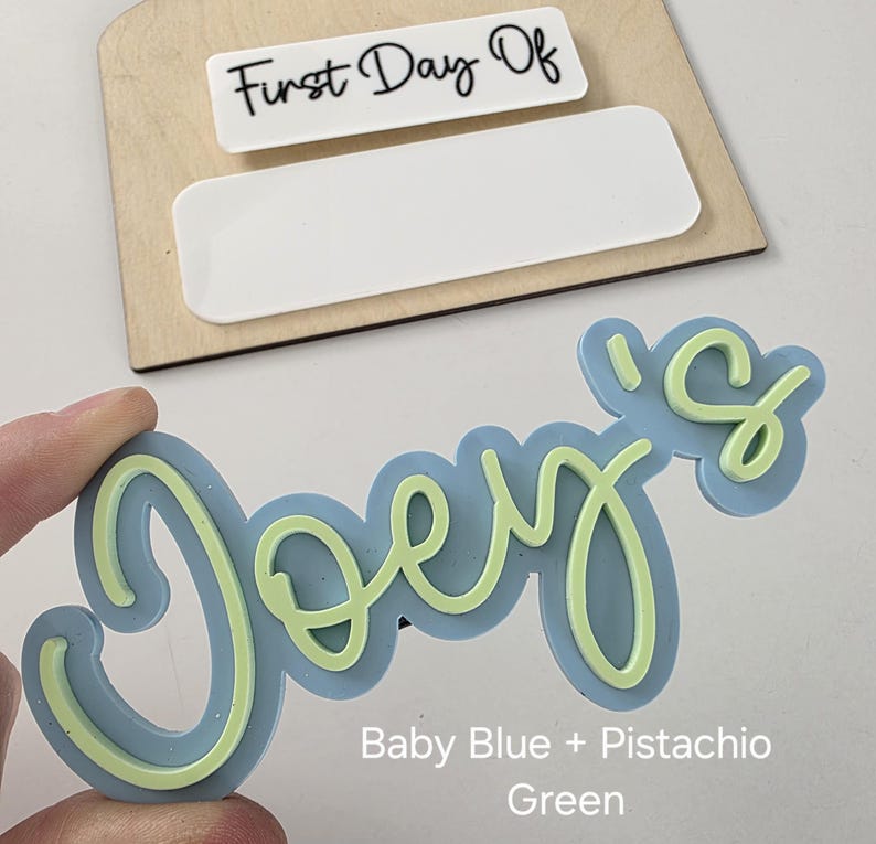 May include: A light blue and pistachio green "Joe's" name sign with a cursive font. The sign is made of wood and has a blank space for a date or other text.