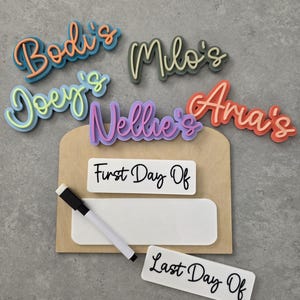 May include: A wooden sign with a white dry-erase board and a black dry-erase marker. The sign has the words "First Day Of" and "Last Day Of" written on it. There are also five colourful name tags that say "Bodi's", "Joey's", "Milo's", "Nellie's", and "Aria's".