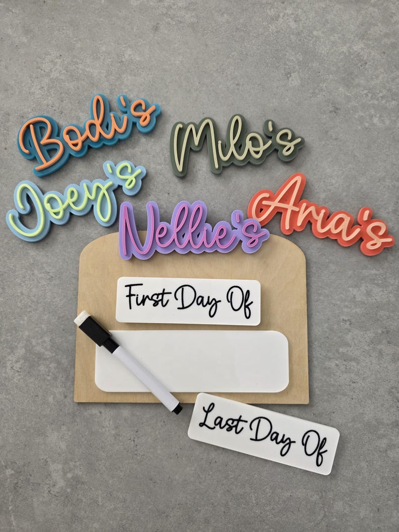 May include: A wooden sign with a white dry-erase board and a black dry-erase marker. The sign has the words "First Day Of" and "Last Day Of" written on it. There are also five colourful name tags that say "Bodi's", "Joey's", "Milo's", "Nellie's", and "Aria's".