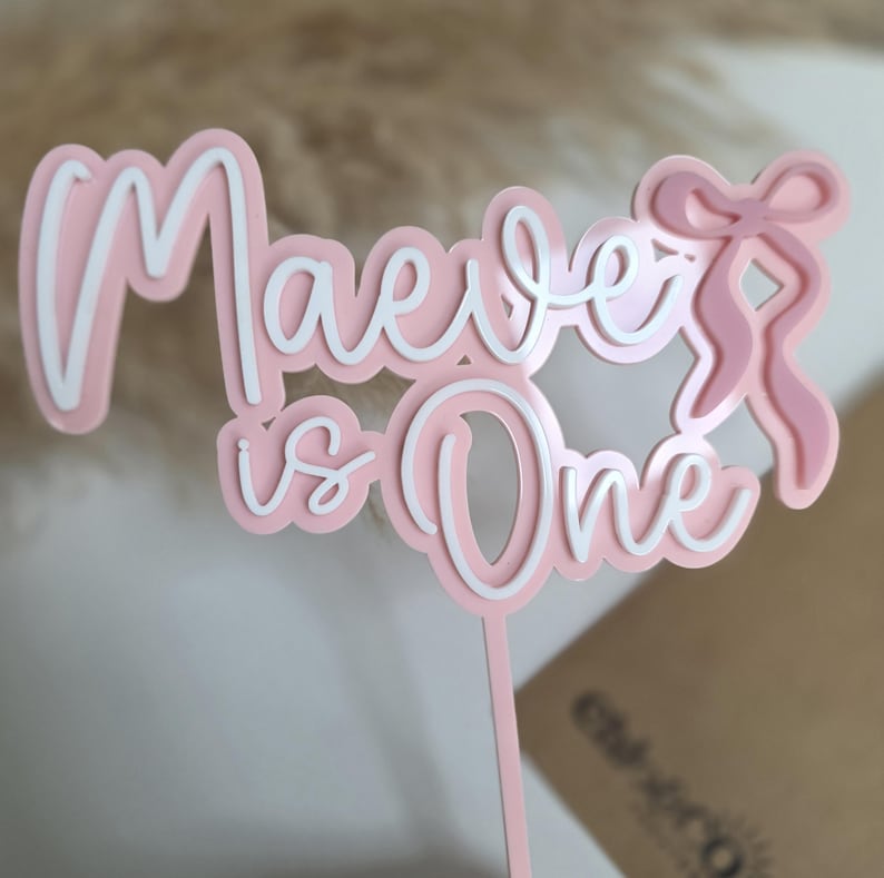 May include: A pink and white cake topper with the words "Maeve is One" in a layered design. The topper includes a bow on the right side and a stem for insertion into a cake. Perfect for a first birthday celebration.