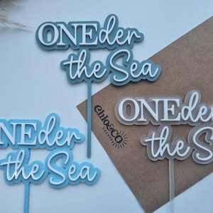 May include: Three cake toppers featuring the text ONEder the Sea in layered acrylic. One topper is dark blue, one is light blue, and one is translucent frosted white. Each topper has a vertical stem for insertion into a cake or food item.