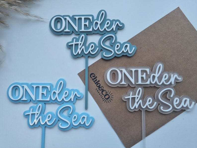 May include: Three cake toppers featuring the text ONEder the Sea in layered acrylic. One topper is dark blue, one is light blue, and one is translucent frosted white. Each topper has a vertical stem for insertion into a cake or food item.