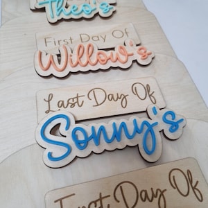 May include: Five wooden signs with the text "Day Of" and a name in a colorful script. The names are Theo's, Willow's, Sonny's, and Olivia's. The signs are stacked on top of each other.