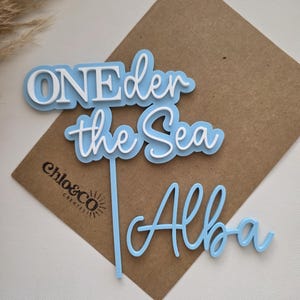 May include: A light blue and white acrylic cake topper featuring the text ONEder the Sea with a vertical stake and a separate light blue script name tag reading Alba. The design uses layered lettering with white text on a light blue background.