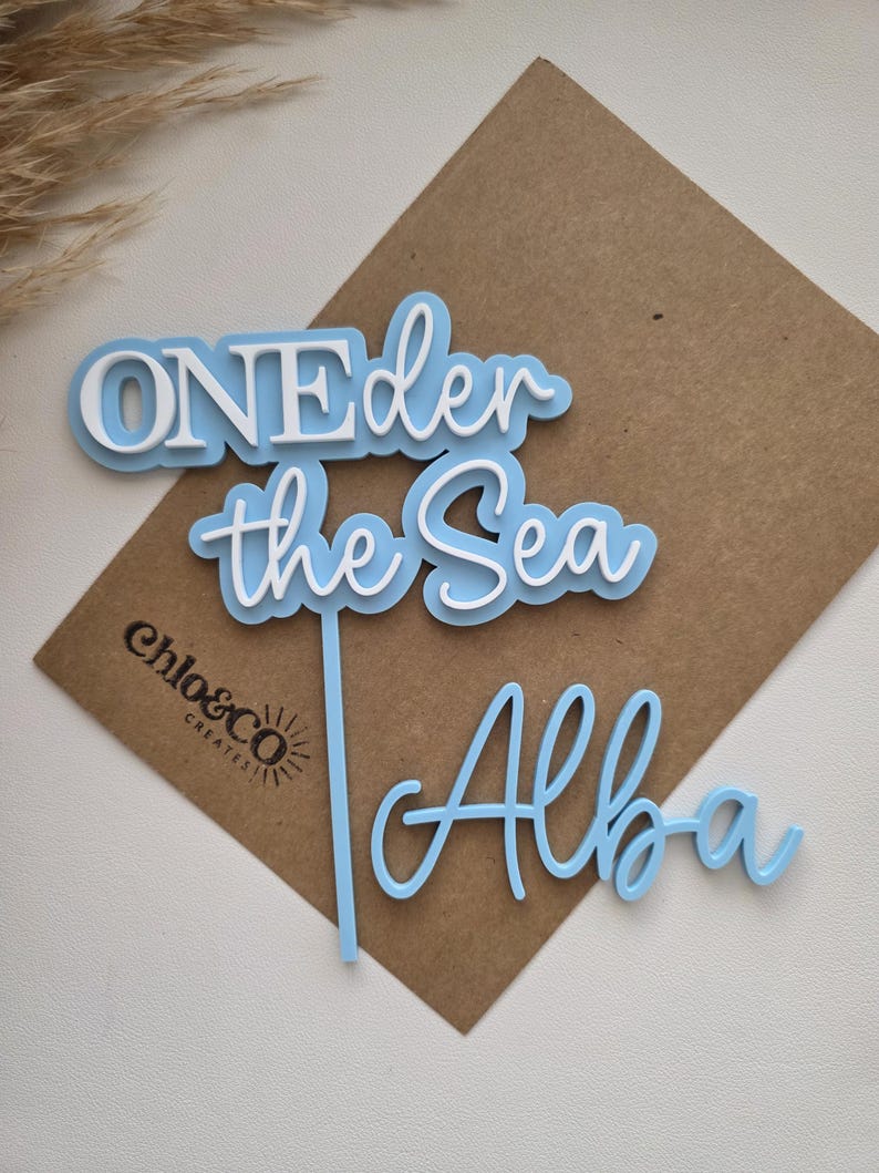 May include: A light blue and white acrylic cake topper featuring the text ONEder the Sea with a vertical stake and a separate light blue script name tag reading Alba. The design uses layered lettering with white text on a light blue background.