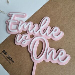 Puede incluir: Topper de pastel de acr&iacute;lico rosa y blanco con el texto "Emilie tiene un a&ntilde;o".