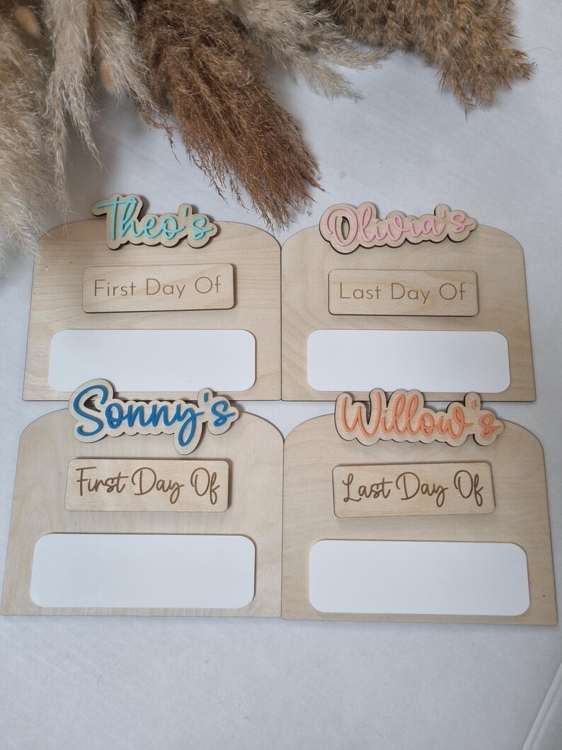 May include: Four wooden photo frames with blank spaces for pictures. Each frame has a name at the top, "Theo's", "Olivia's", "Sonny's", and "Willow's", and a sign that says "First Day Of" or "Last Day Of".