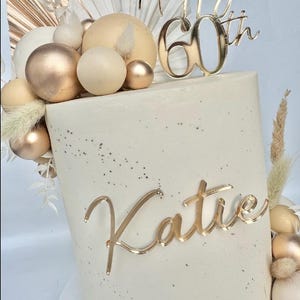 May include: A white cake decorated with gold and beige round ornaments, a gold 'Happy 60th' topper, and a gold 'Katie' topper.
