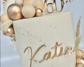 Puede incluir: Un pastel blanco decorado con adornos redondos dorados y beige, un adorno dorado 'Happy 60th' y un adorno dorado 'Katie'.