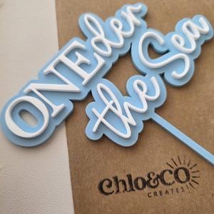 May include: A light blue and white cake topper featuring the text ONEDER the Sea in a layered design. The topper has a light blue base layer with white raised lettering and a thin light blue stick extending from the bottom right side.