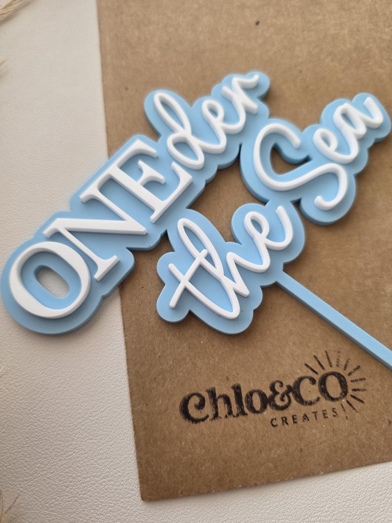 May include: A light blue and white cake topper featuring the text ONEDER the Sea in a layered design. The topper has a light blue base layer with white raised lettering and a thin light blue stick extending from the bottom right side.