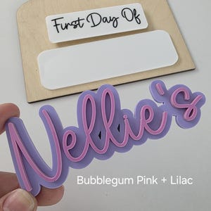 May include: A lilac and bubblegum pink acrylic name sign that says "Nellie's".
