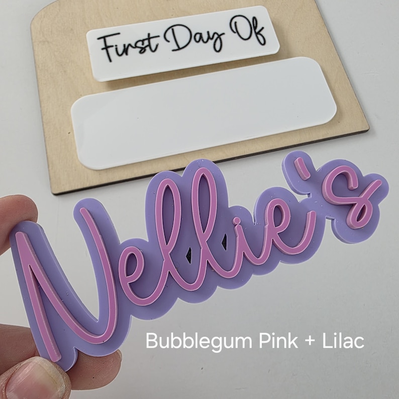 May include: A lilac and bubblegum pink acrylic name sign that says "Nellie's".