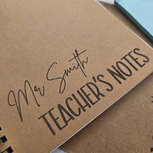 May include: A spiral-bound notebook with a kraft paper cover. The cover is printed with the words "Mr Smith TEACHER'S NOTES" in black ink. A pen rests on the notebook. A light blue sticky note pad is in the background.