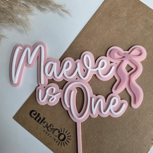 May include: Pink and white cake topper with the text "Maeve is One" in a cursive font. The topper has a bow design and a stick for insertion into a cake. The logo "chloe&co creates" is visible.