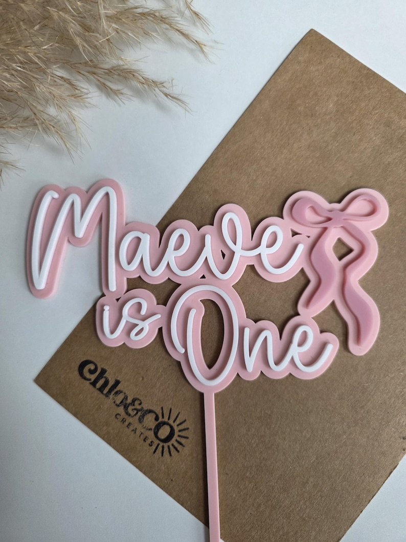 May include: Pink and white cake topper with the text "Maeve is One" in a cursive font. The topper has a bow design and a stick for insertion into a cake. The logo "chloe&co creates" is visible.