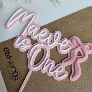 May include: A pink and white cake topper with the words "Maeve is One" in a script font. The topper includes a bow design and a stem for insertion into a cake. The logo "chlo&co creates" is visible.