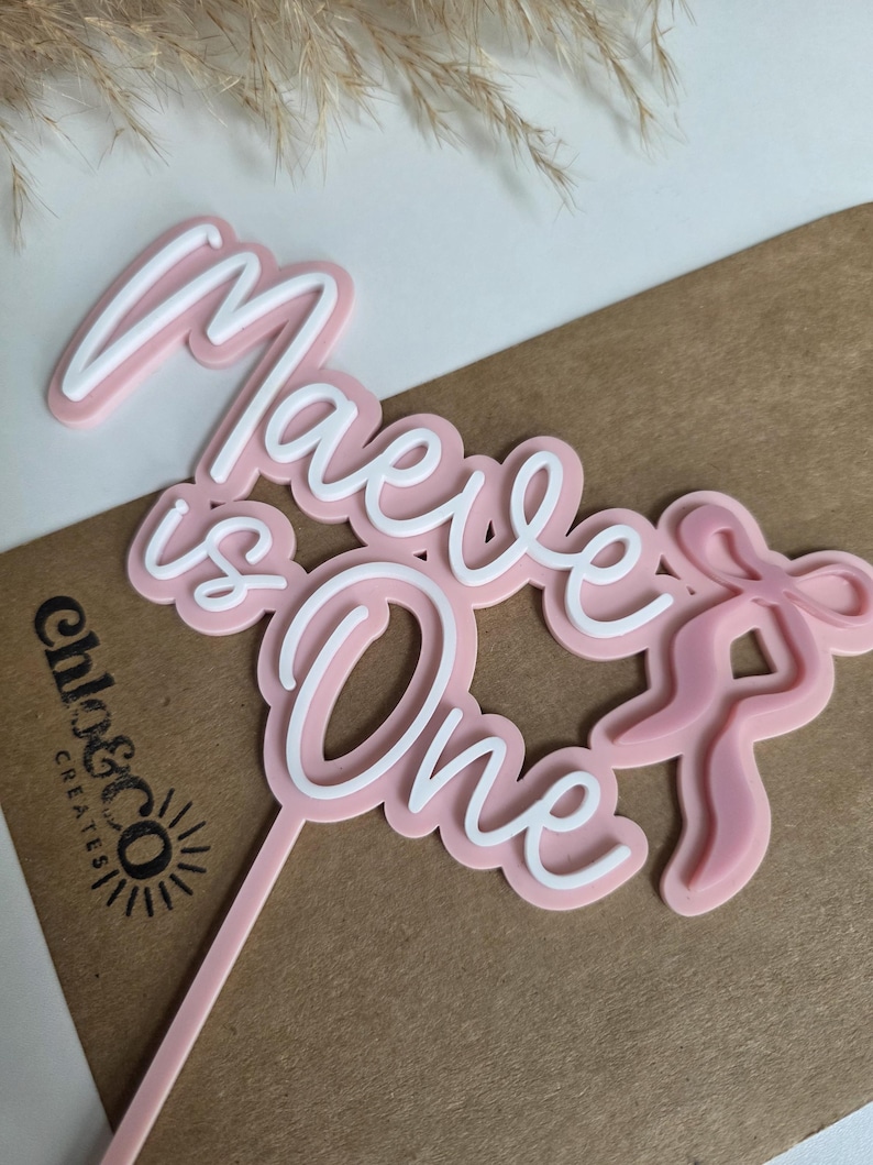 May include: A pink and white cake topper with the words "Maeve is One" in a script font. The topper includes a bow design and a stem for insertion into a cake. The logo "chlo&co creates" is visible.