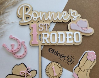 Könnte beinhalten: Ein Kuchenaufsatz aus Holz mit dem Text "Bonnie's 1st Rodeo" in Weiß und Rosa. Das Bild enthält ein Hufeisen, einen Cowboyhut, einen Stiefel und ein Band mit der Nummer 1. Für eine Geburtstagsparty im Rodeo-Stil.