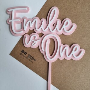 Puede incluir: Topper de pastel de acr&iacute;lico rosa y blanco con el texto "Emilie is One" en una fuente cursiva. El topper est&aacute; en un palo rosa.