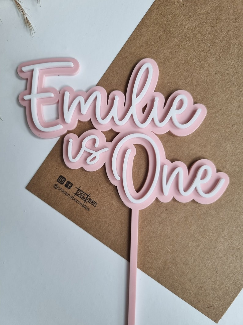 Puede incluir: Topper de pastel de acr&iacute;lico rosa y blanco con el texto "Emilie is One" en una fuente cursiva. El topper est&aacute; en un palo rosa.