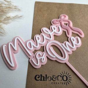 May include: A pink and white cake topper with the words "Maeve is One" in a cursive font. The topper has a bow design and a stick for insertion into a cake. The logo "chloe&co CREATES" is visible.