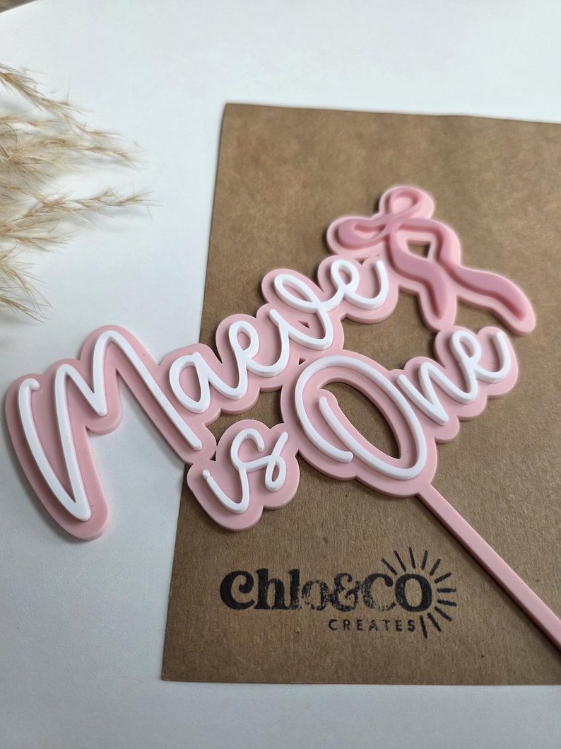 May include: A pink and white cake topper with the words "Maeve is One" in a cursive font. The topper has a bow design and a stick for insertion into a cake. The logo "chloe&co CREATES" is visible.