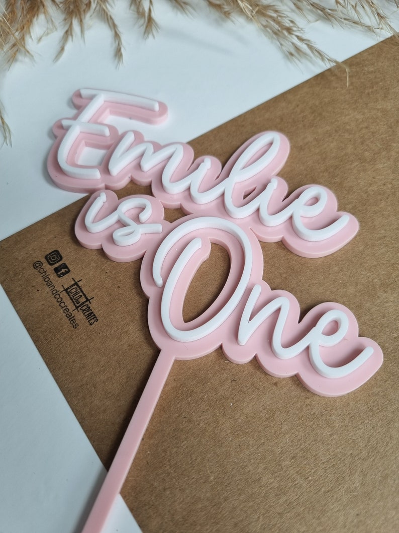 Puede incluir: Topper de pastel de acr&iacute;lico rosa con las palabras "Emilie is One" en letras blancas. El topper est&aacute; sobre una superficie marr&oacute;n.