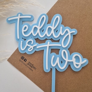 May include: Blue and white acrylic cake topper with the text "Teddy is Two".