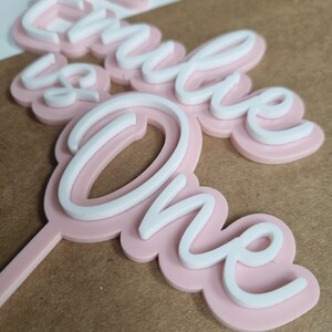 Puede incluir: Topper de pastel rosa y blanco con las palabras "Hello One" en una fuente cursiva.