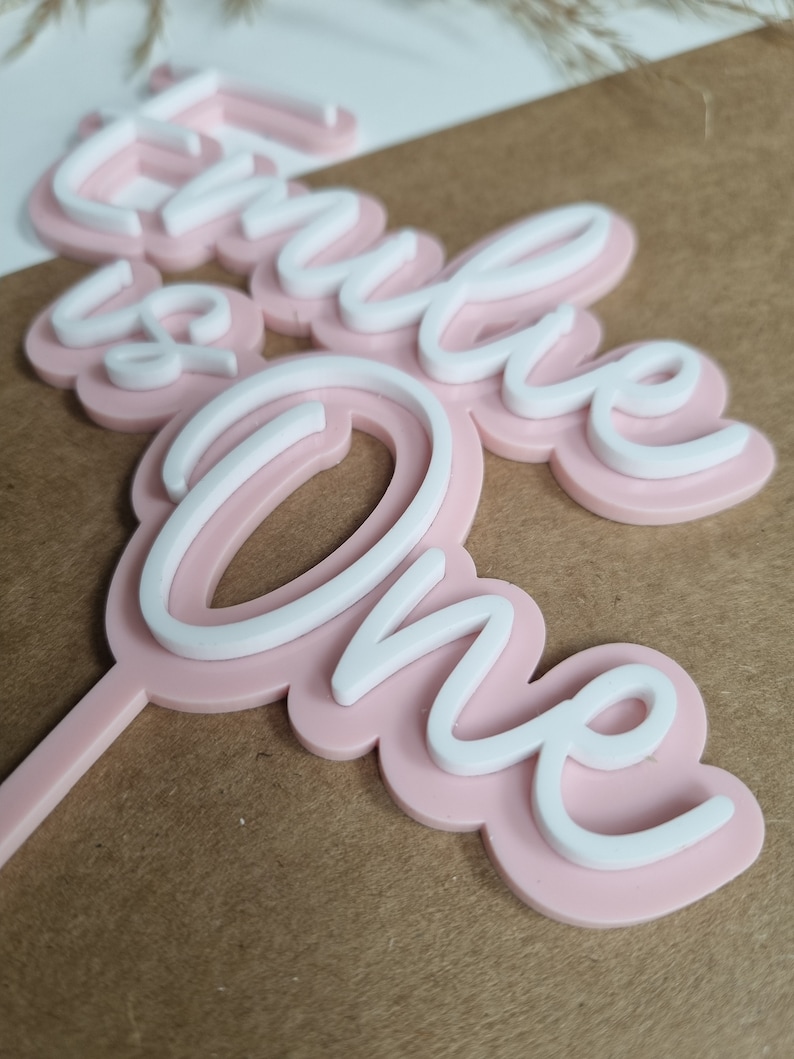 Puede incluir: Topper de pastel rosa y blanco con las palabras "Hello One" en una fuente cursiva.