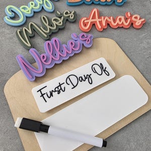 May include: A wooden board with a white dry-erase surface and a black marker. The board has the words "First Day Of" written on it. There are also five colourful, 3D-printed name tags with the names "Doey's", "Bodi's", "Milo's", "Anais", and "Nellie's" on them.