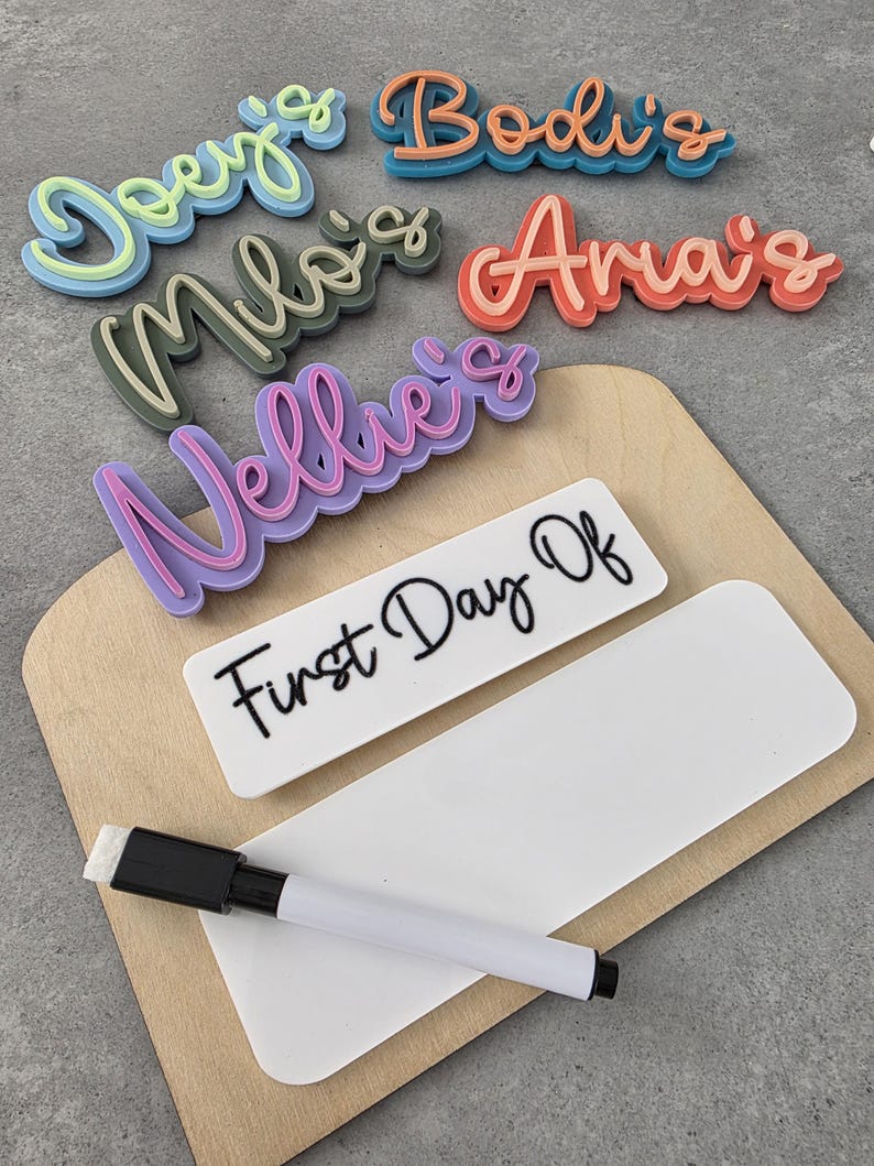 May include: A wooden board with a white dry-erase surface and a black marker. The board has the words "First Day Of" written on it. There are also five colourful, 3D-printed name tags with the names "Doey's", "Bodi's", "Milo's", "Anais", and "Nellie's" on them.