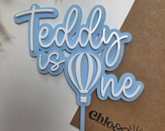 Könnte beinhalten: Blau-weiße Tortenaufsatz mit dem Schriftzug "Teddy is One" und einem Heißluftballon-Design. Der Aufsatz ist zum Einstecken in einen Kuchen gedacht. Das Logo "chlo&co CREATES" ist sichtbar.